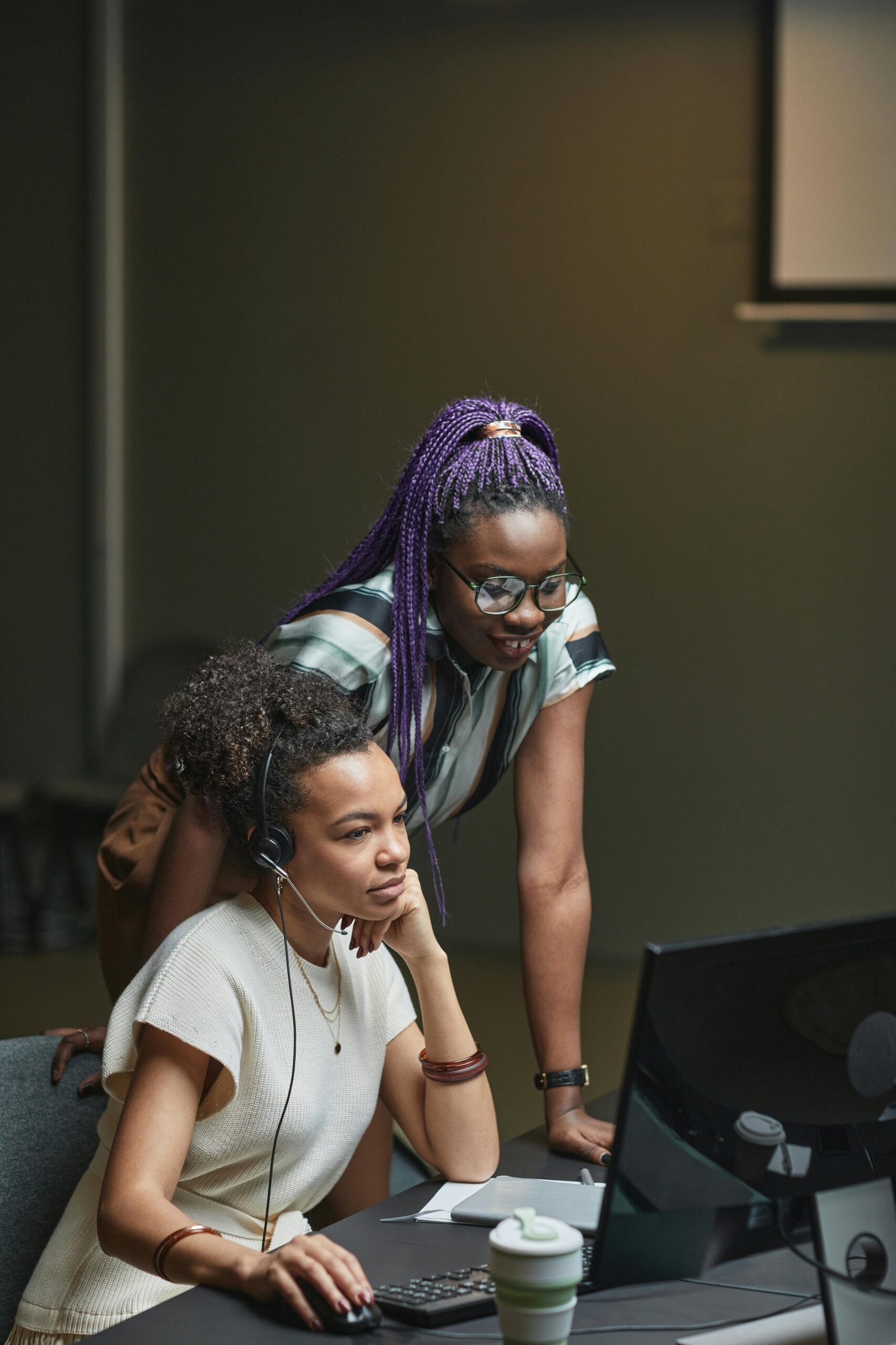 Two women working together at an office desk, focusing on a computer screen.